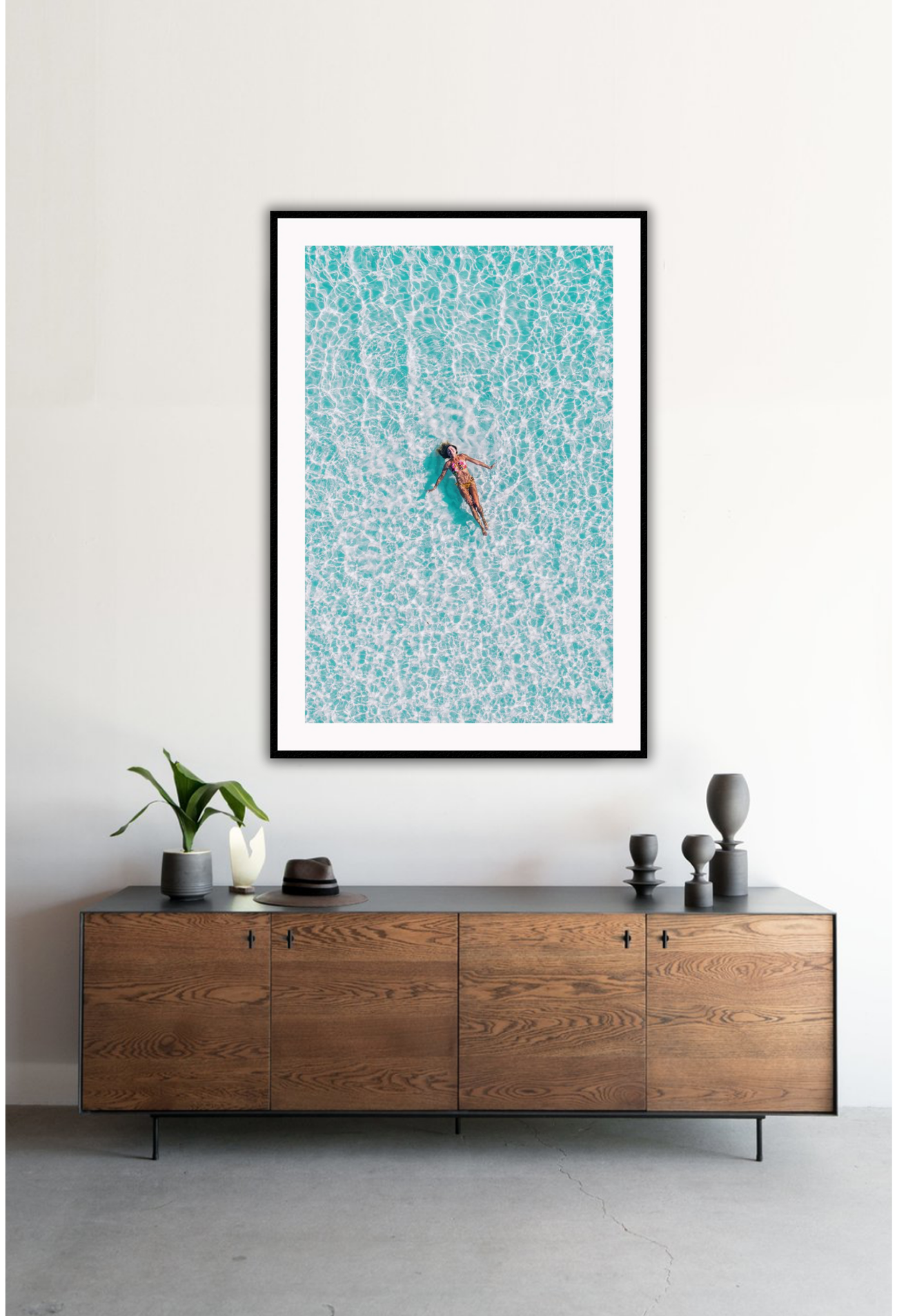 A framed print depicting a person swimming in a turquoise ocean, with a high quality custom frame. Hung on a white wall above a timber cabinet.