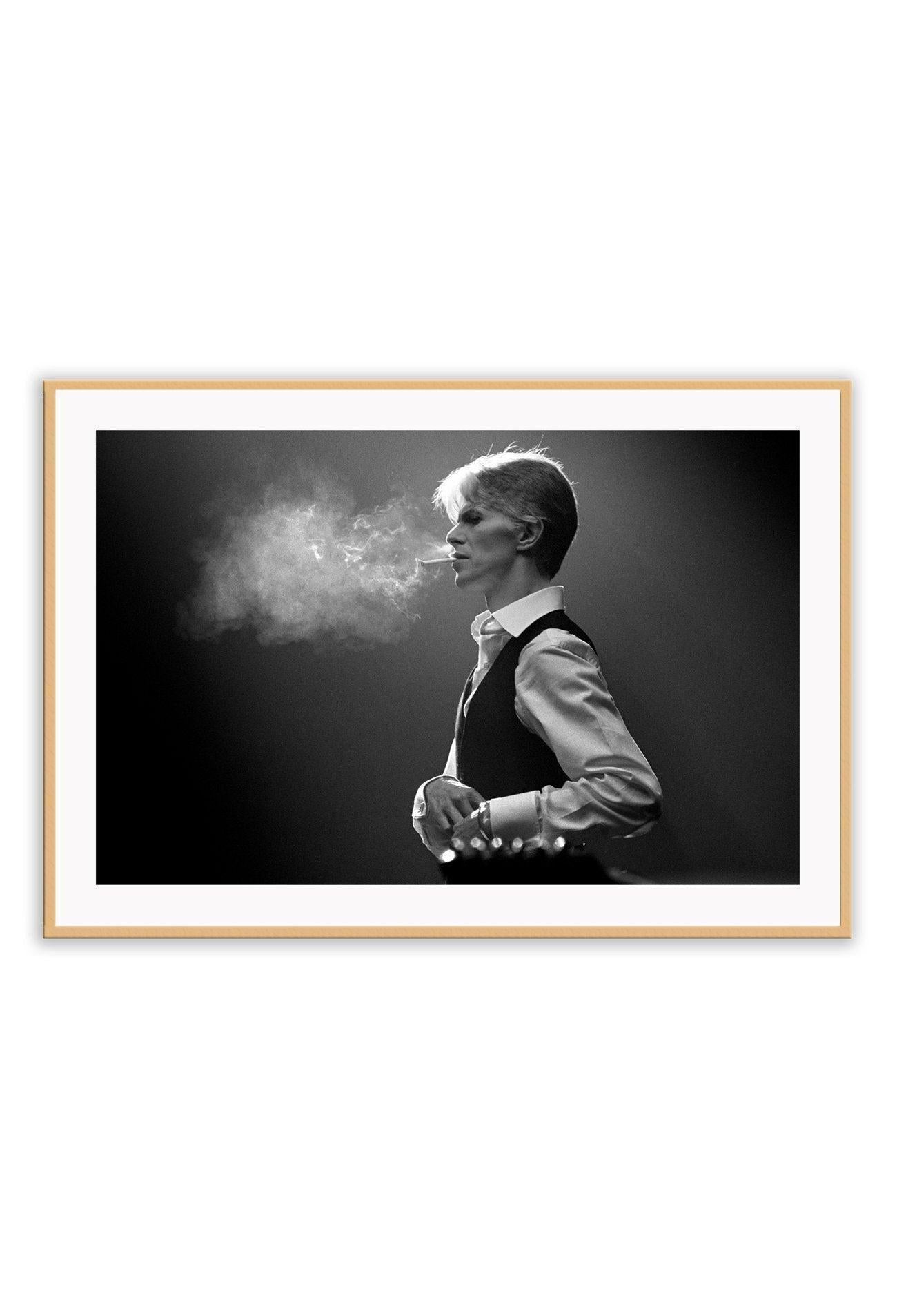 A framed black and white photograph of David Bowie, with smoke surrounding him, mounted in a frame with a matte wood finish.