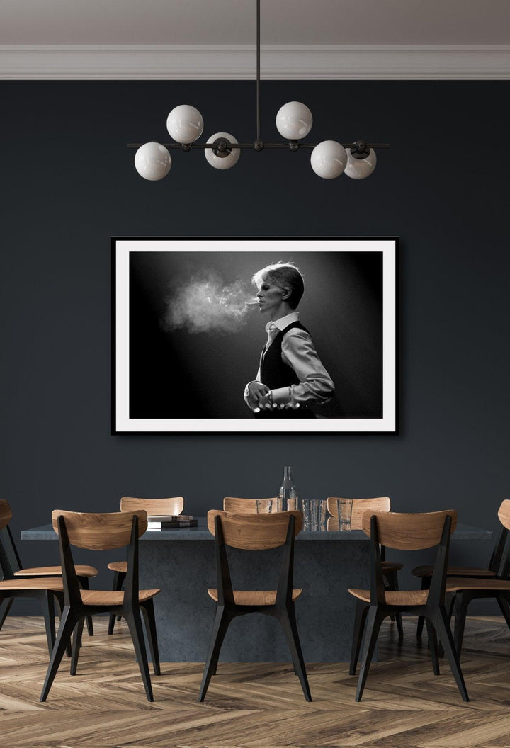 A framed black and white photograph of David Bowie,  smoking, mounted in a frame with a matte wood finish on a dark grey wall hanging over a dining room table.